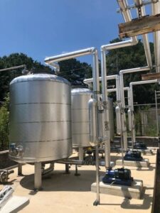 A row of metallic industrial storage tanks with connected piping and pumps in a sunny outdoor setting.